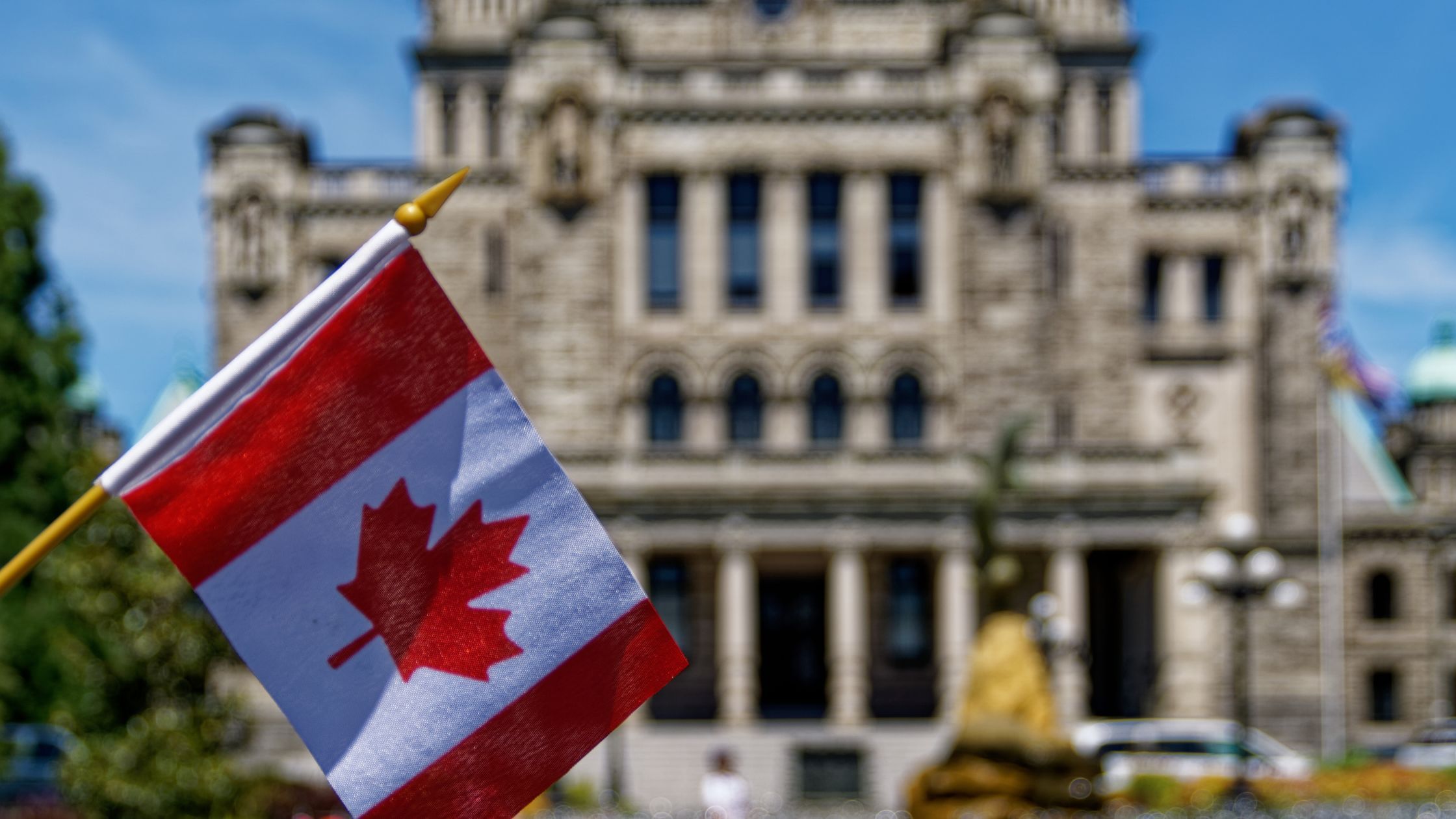 A Canadian flag in focus with a blurred background of a historic stone building and trees under a clear sky.
